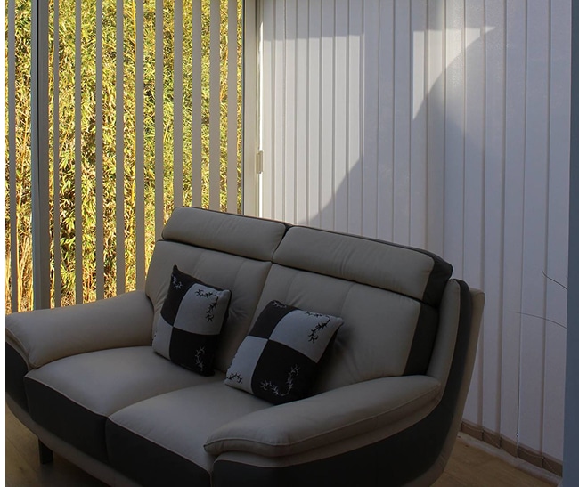 Stores Verticaux Salon Canapé Lumière Ambiante Canapé moderne beige et gris devant de grandes fenêtres équipées de stores verticaux clairs filtrant la lumière du soleil et la verdure extérieure.