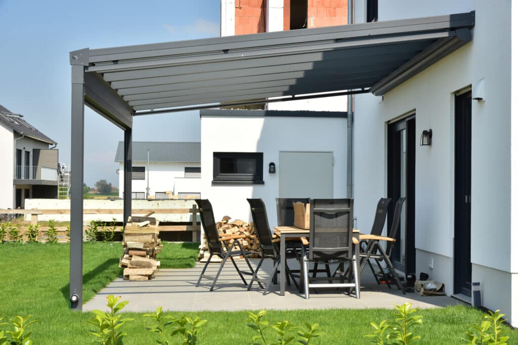 Patio Moderne : Pergola Bioclimatique et Mobilier Extérieur Patio moderne avec pergola bioclimatique grise, table et chaises noires, adossée à une maison blanche en construction, avec des bûches et pelouse verte.