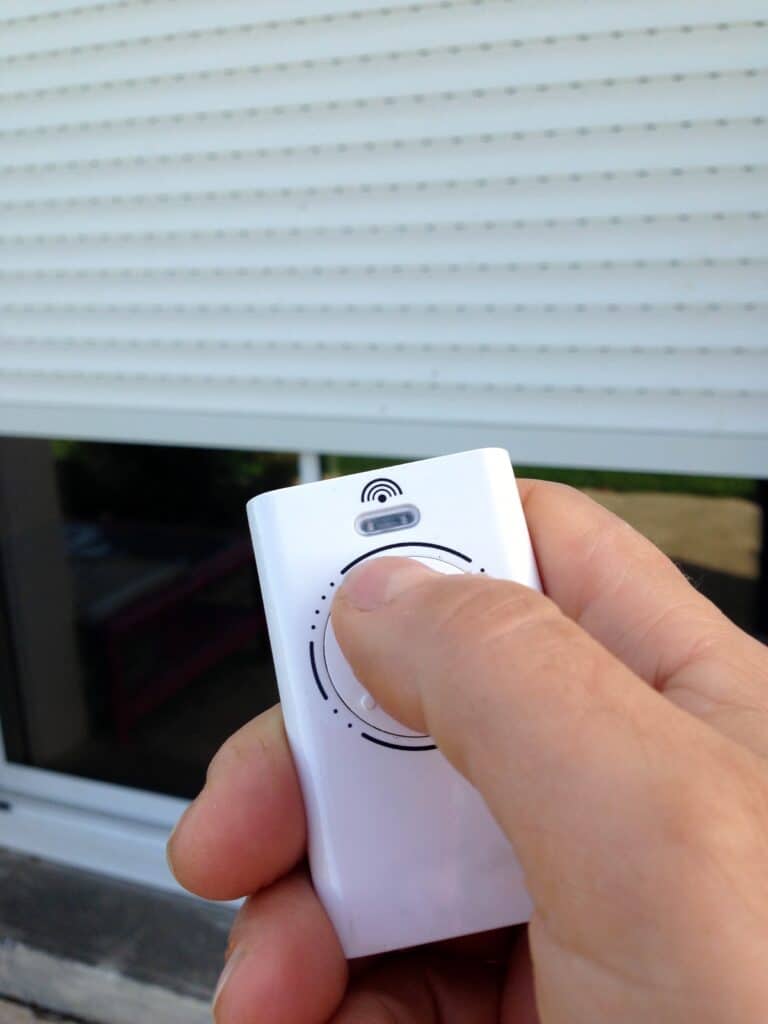 Télécommande sans fil pour volet roulant : contrôle facile Main actionnant une télécommande blanche pour volet roulant motorisé, avec un voyant et un cadran de contrôle visibles.