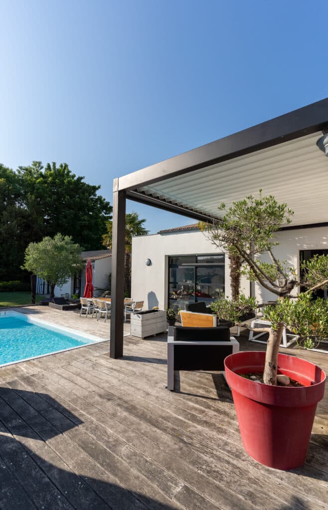 Pergola Moderne, Terrasse Bois et Piscine Lumineuse Terrasse en bois près d'une piscine et d'une maison moderne avec pergola bioclimatique, mobilier extérieur et grand pot rouge.